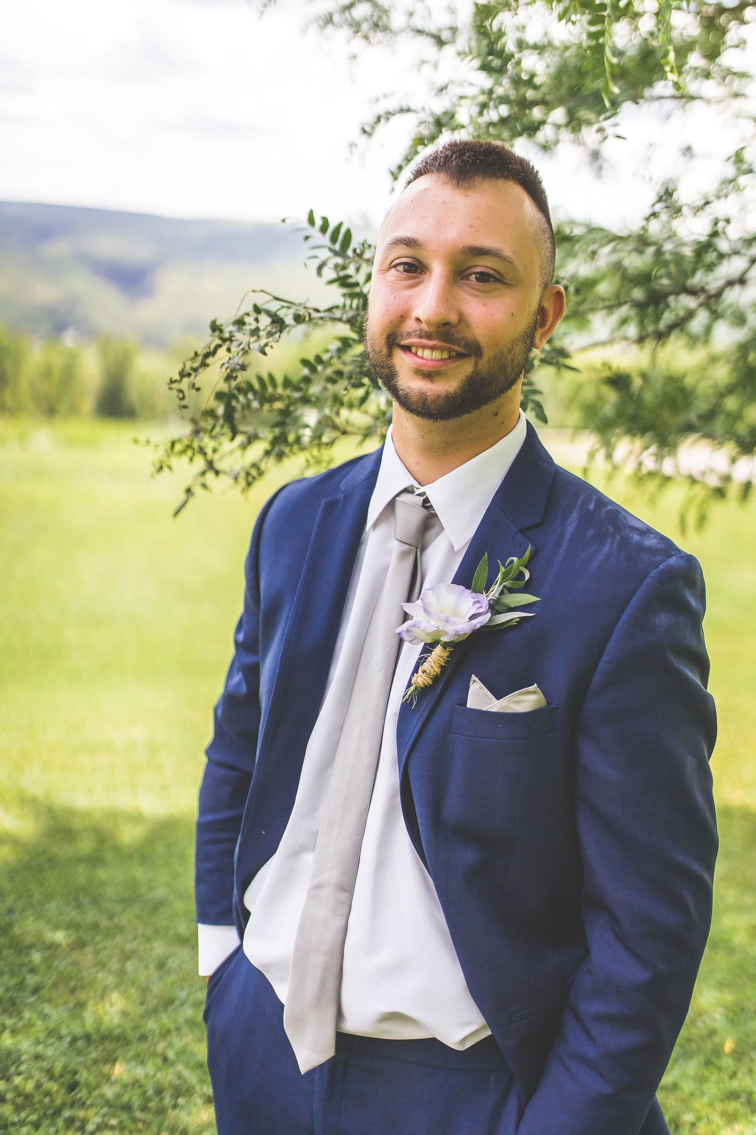 Groom in navy blue suit poses outdoors in a garden setting with natural greenery in the background.