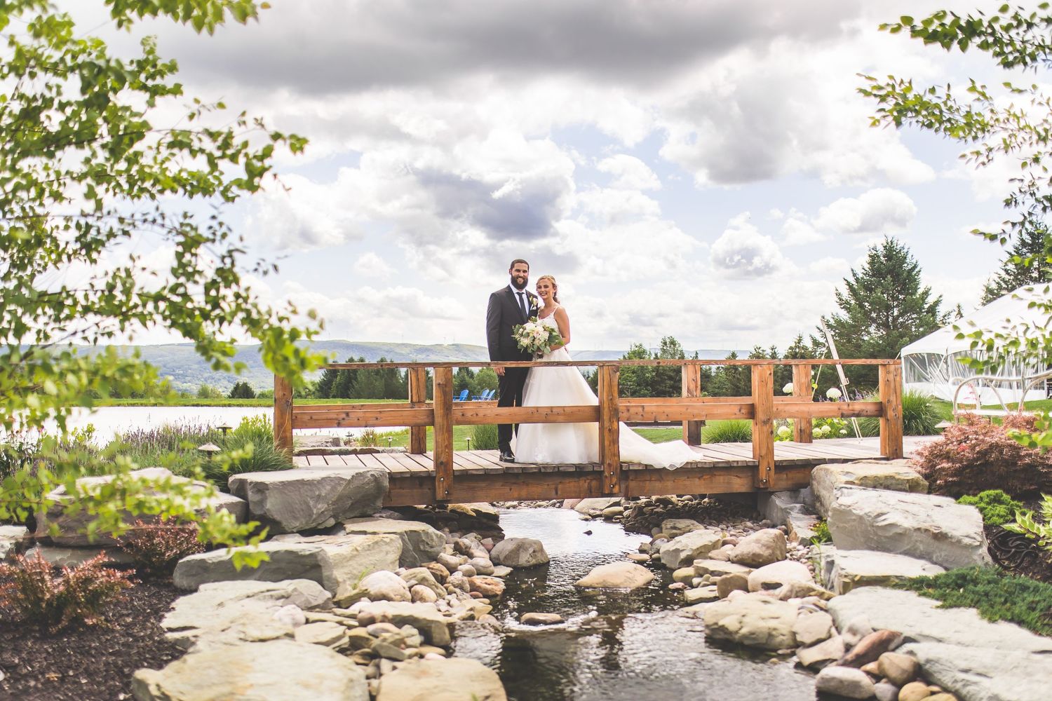 Wedding couple stands on rustic wooden bridge over decorative rock garden.