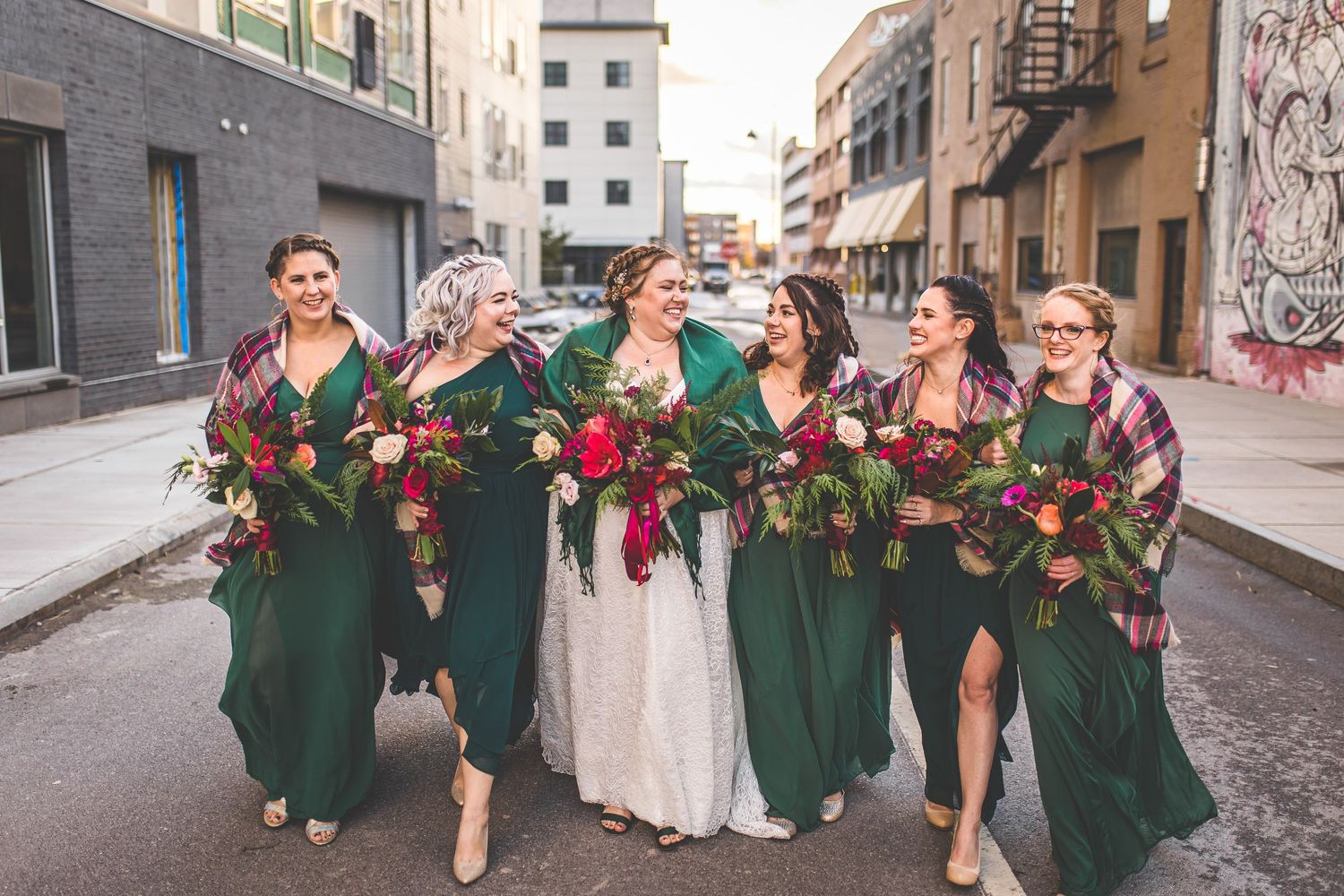 Wedding party members in emerald green dresses and navy suits pose together on an urban street.