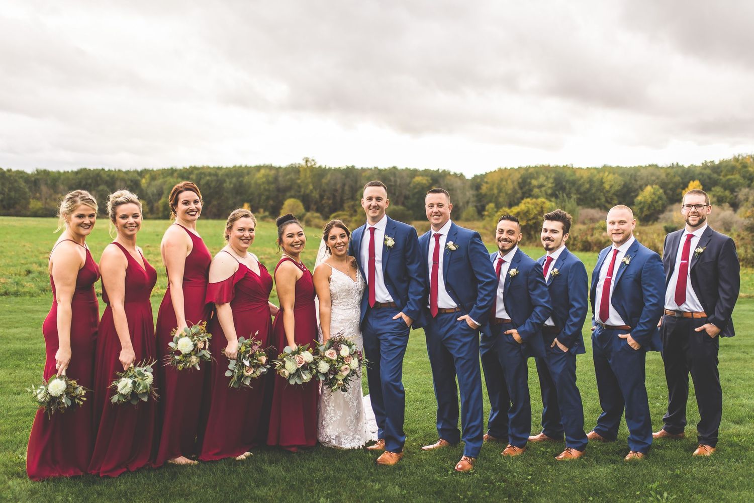 Outdoor wedding party photo with bridesmaids in burgundy dresses and groomsmen in navy suits.