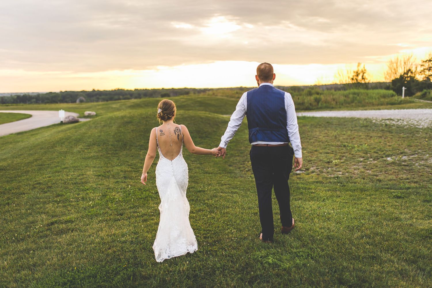 Wedding couple walks hand in hand through a grassy field during sunset.