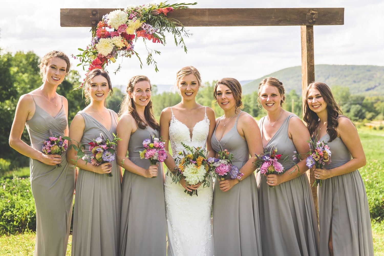 Bridesmaids in gray dresses pose with bouquets under wooden ceremony arch.