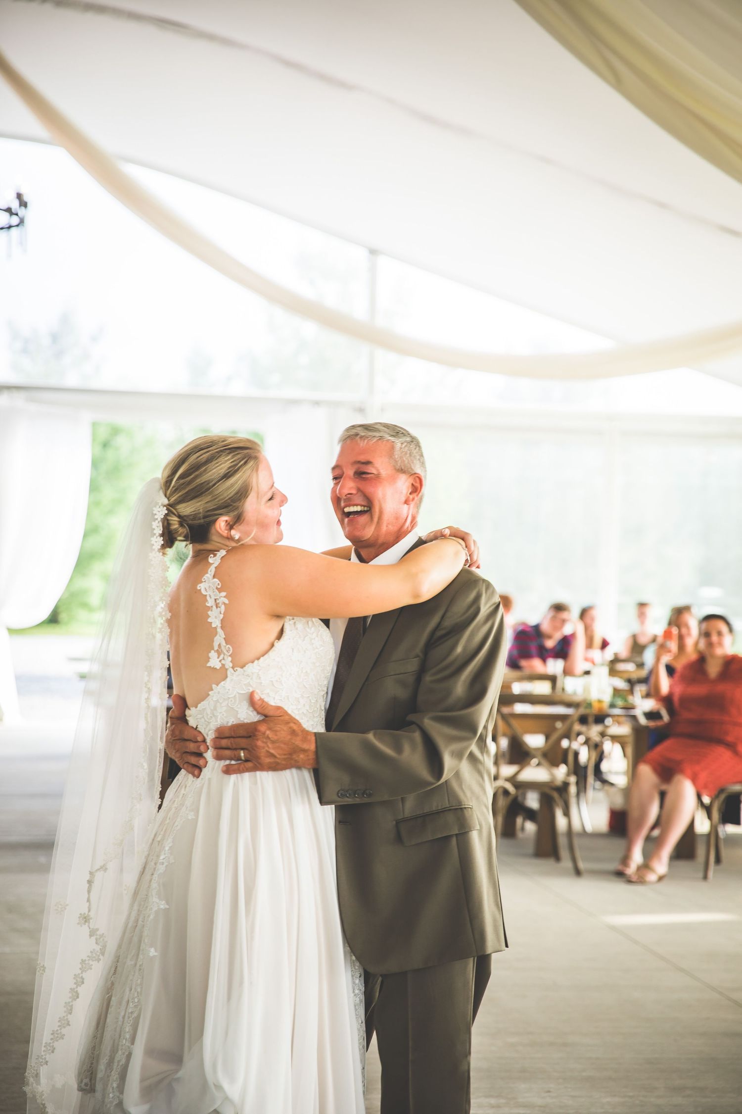 A tender moment during a wedding dance under a draped white tent with guests seated in background.