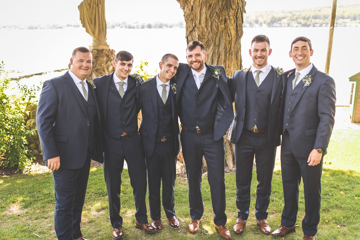 Groomsmen in dark suits pose together at outdoor waterfront wedding venue.
