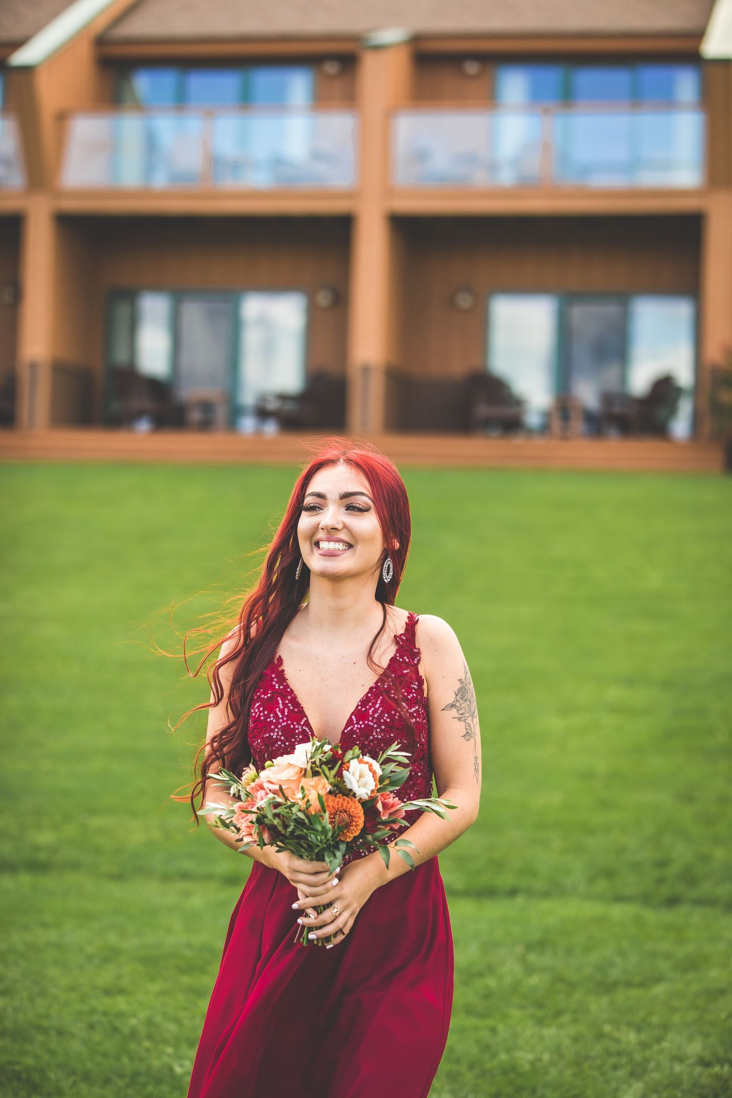 Woman in burgundy dress holding floral bouquet stands on lawn in front of modern building.