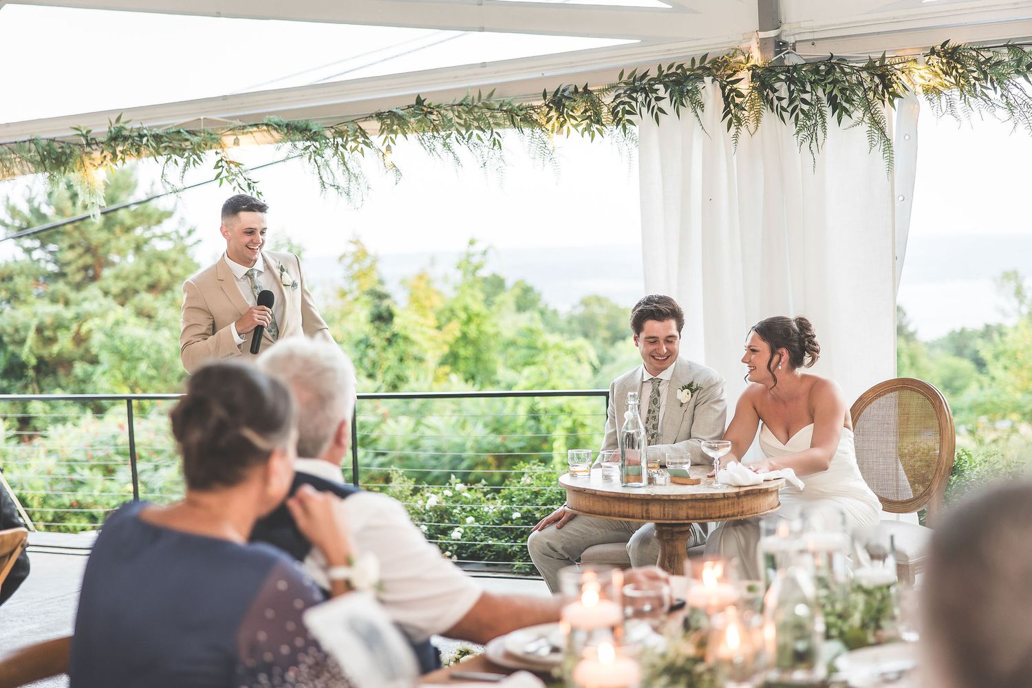 Wedding guests gather under an outdoor tent decorated with greenery and string lights during a celebration.