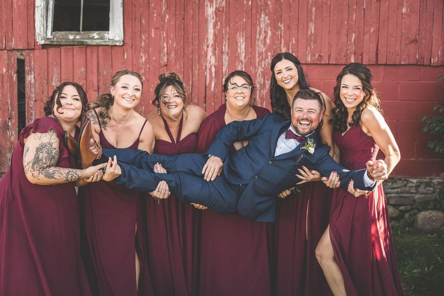 Wedding party members arrange for creative group photos against vintage barn exterior.