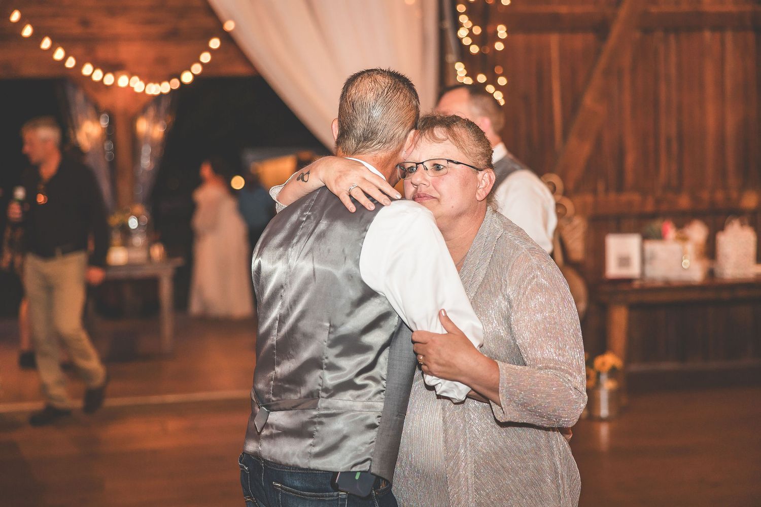 Couples slow dancing at a rustic barn wedding reception with warm string lights overhead.