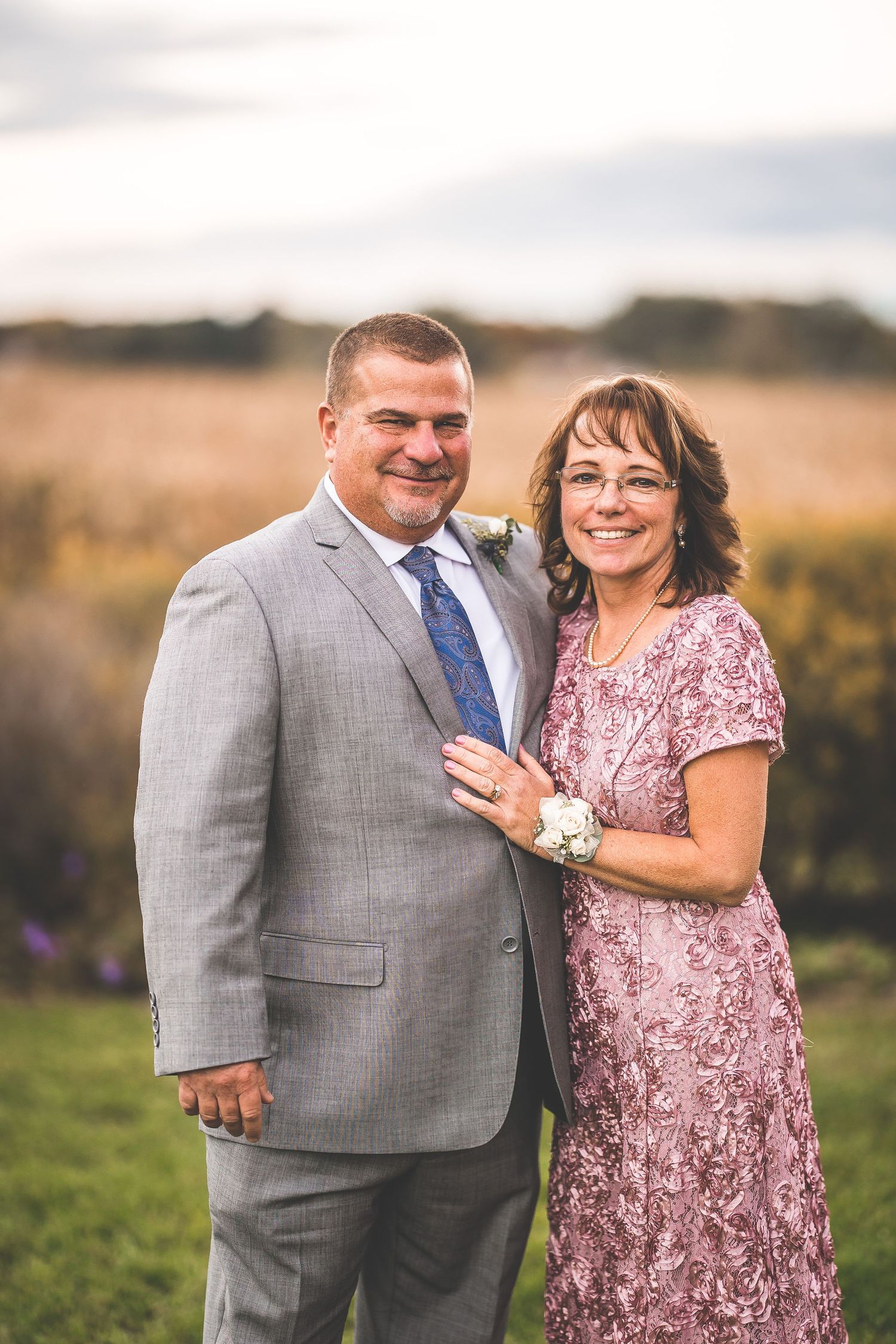 A couple smiles together outdoors wearing pink and grey formal attire.