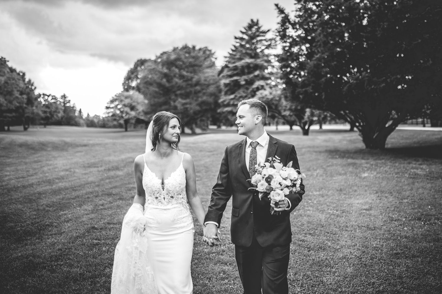Wedding couple walking together on a grassy lawn surrounded by tall pine trees.