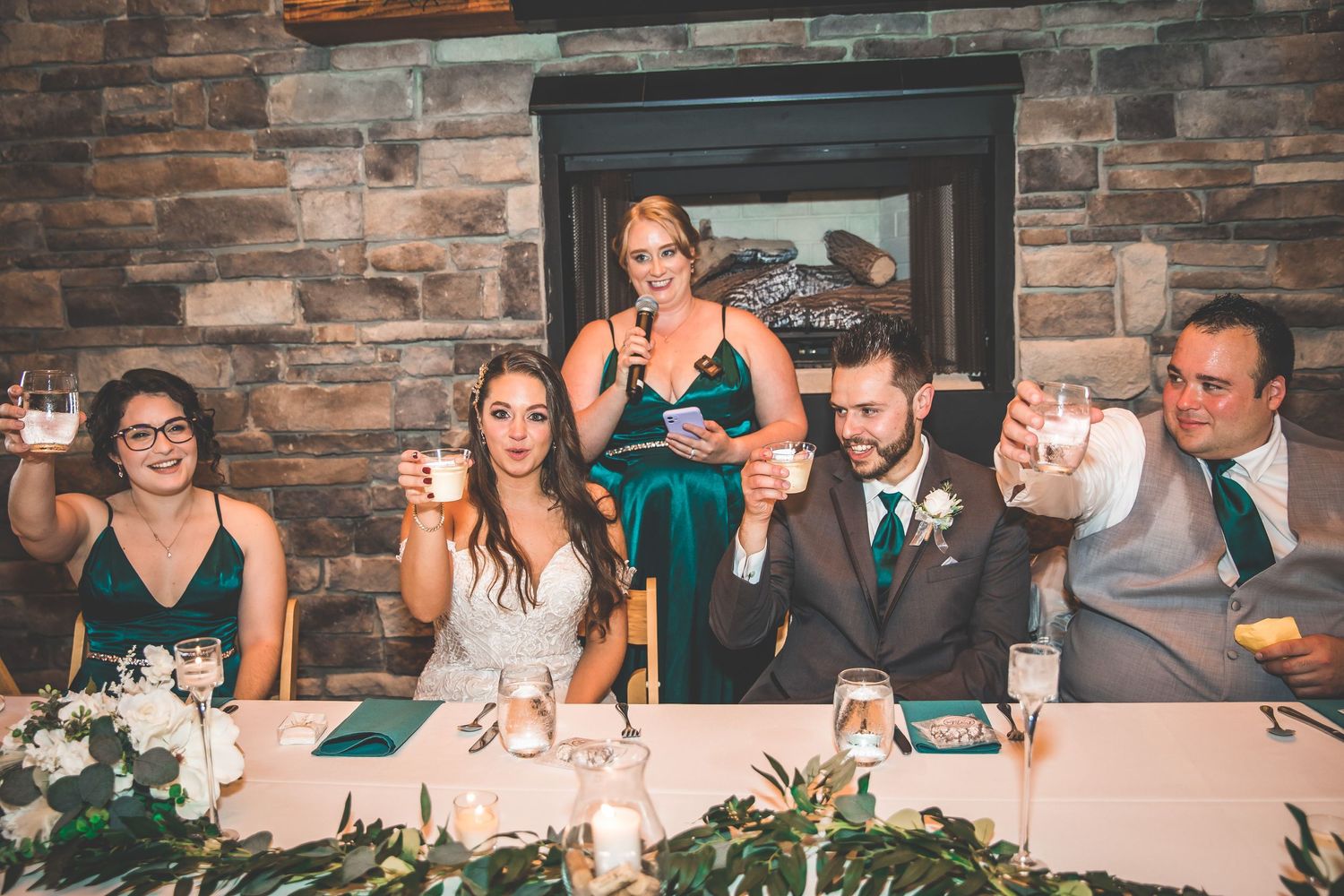 Wedding party members seated at long reception table with rustic decor.