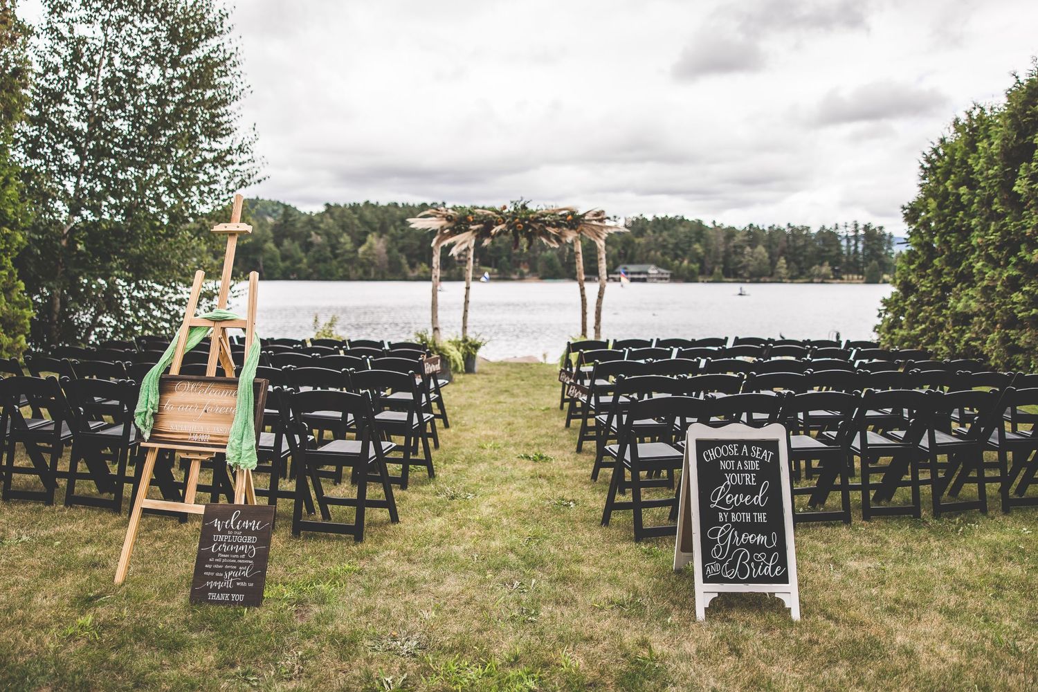 Outdoor wedding ceremony setup with black chairs and wooden arch overlooking a lake.