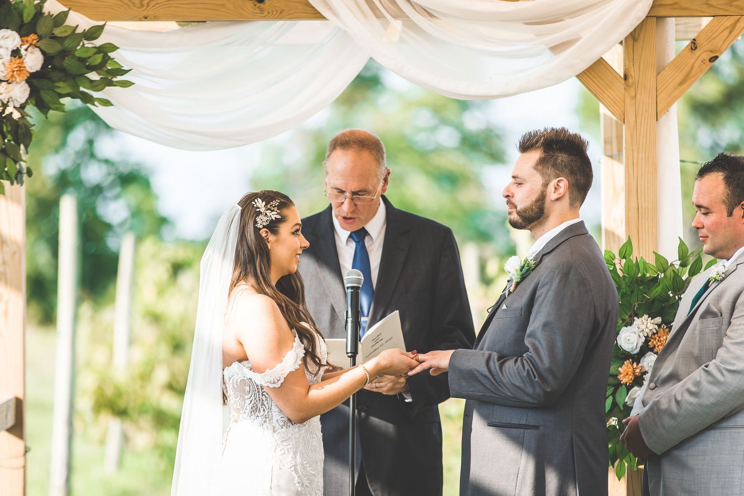 Newlyweds sharing first kiss under wooden pergola decorated with white drapes and flowers.