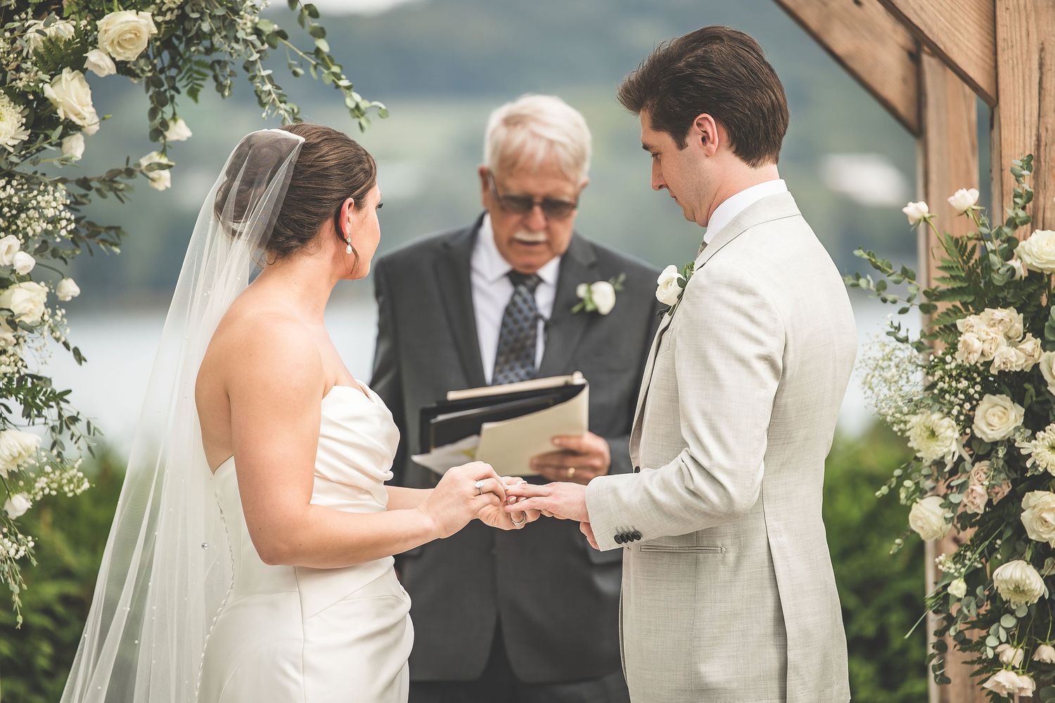 Wedding ceremony exchange of rings under wooden arch decorated with white flowers.
