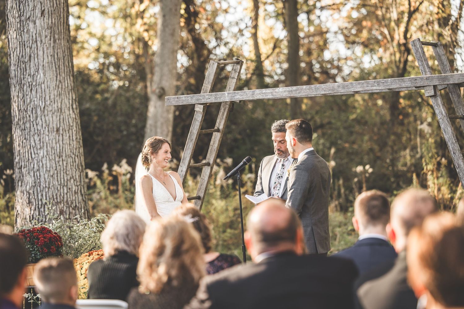 Wedding ceremony taking place under rustic wooden arch in sunlit forest setting.