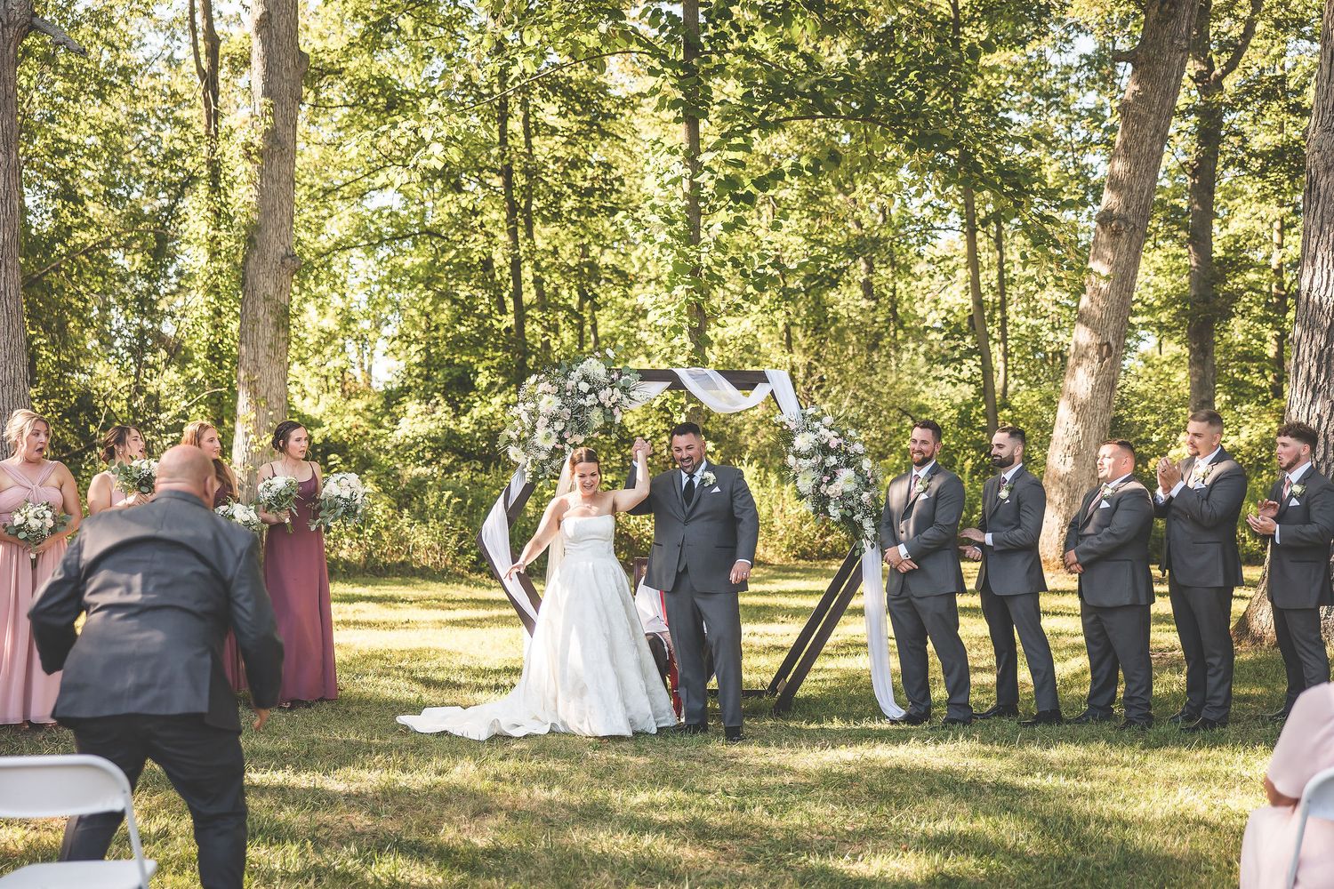 Wedding guests gather for an outdoor ceremony in a sunlit wooded area with white folding chairs.