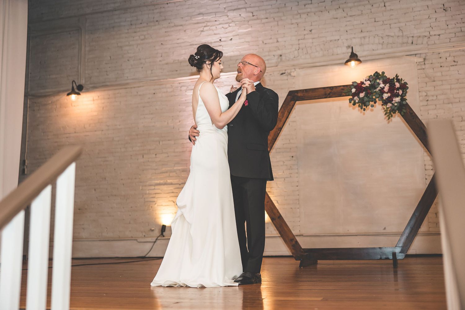 A tender first dance moment captured beneath a geometric wooden arch in an industrial venue.