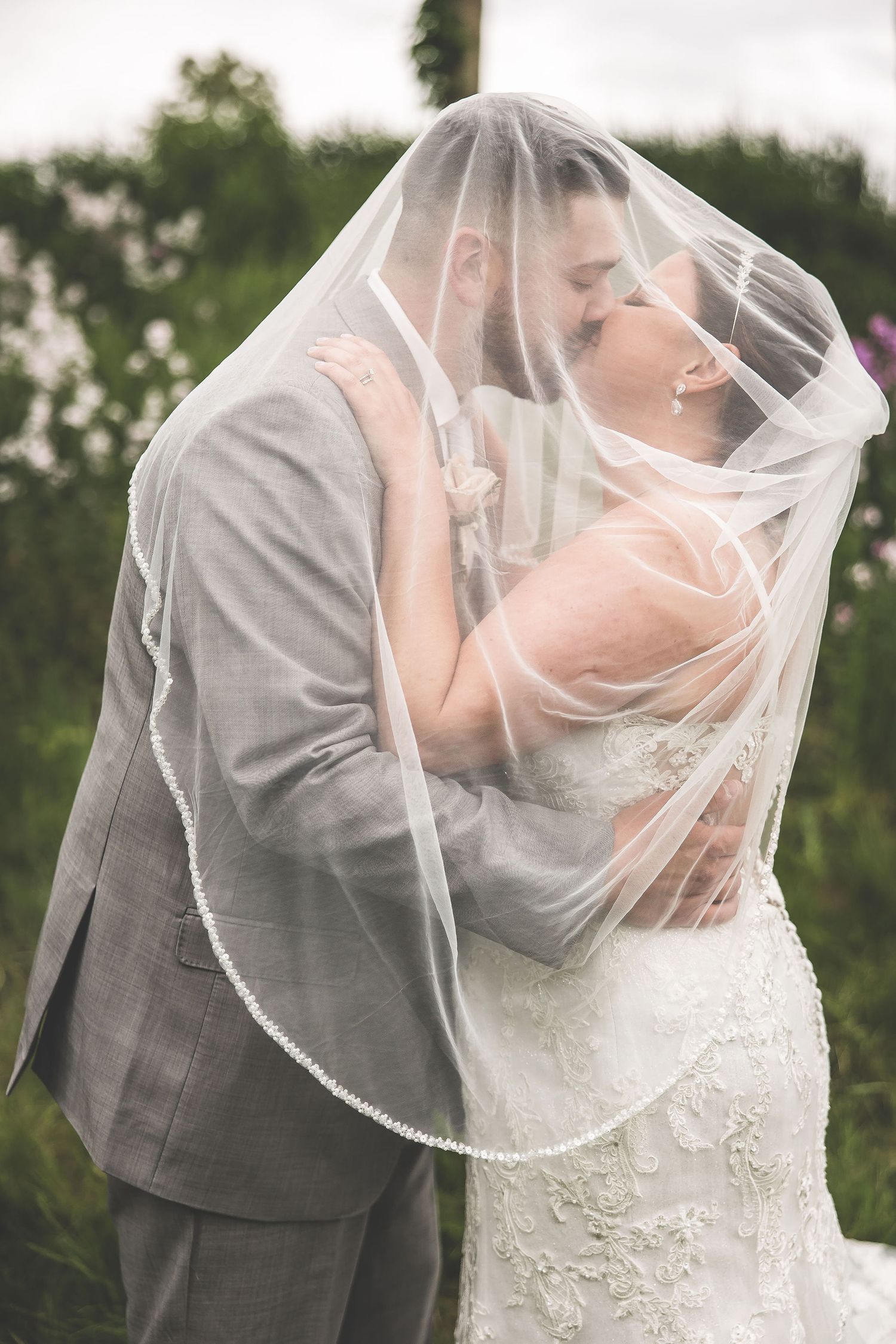 A tender wedding moment captured through a delicate lace veil against a natural outdoor backdrop.