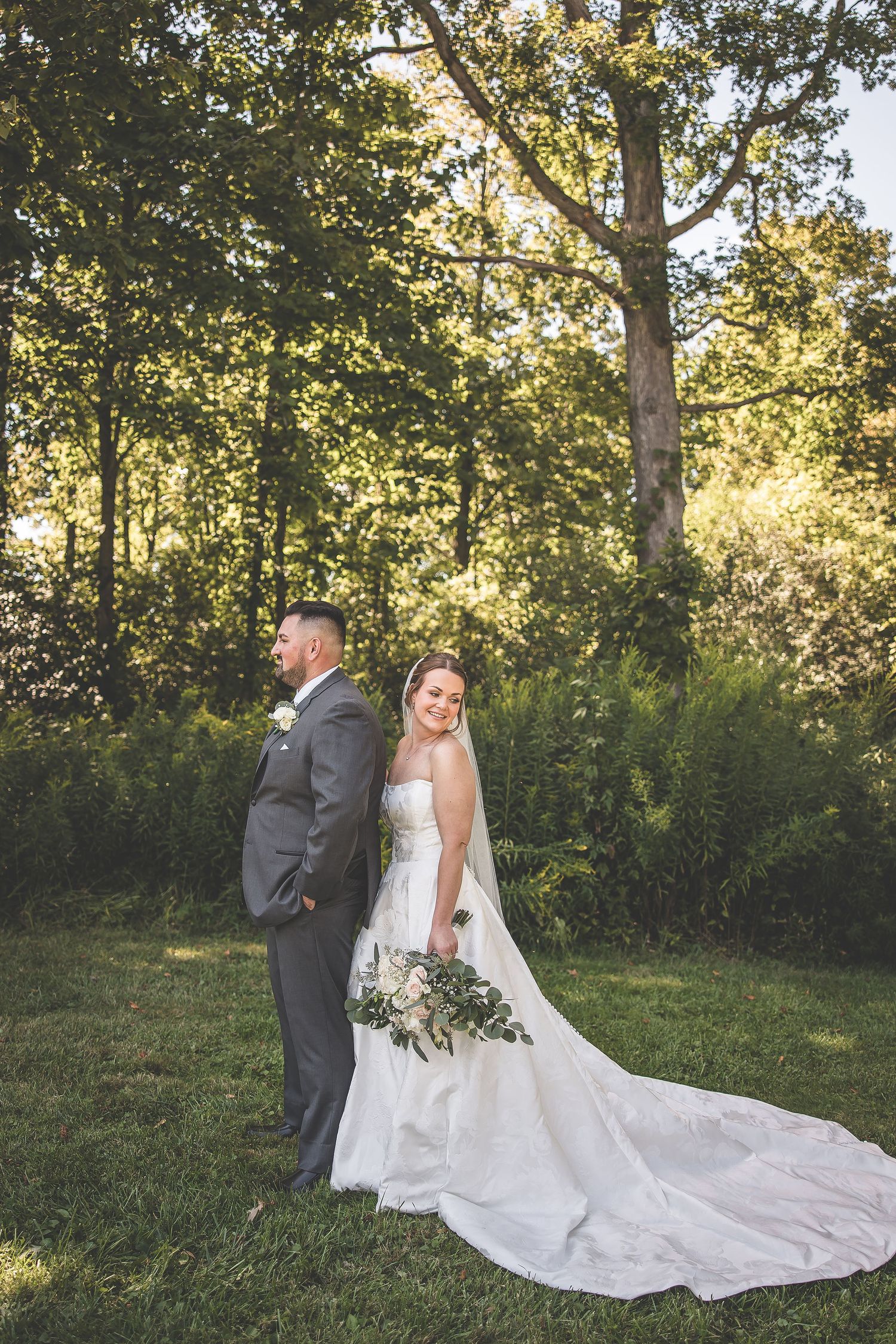 Couple poses for first look wedding photo with long train against woodland backdrop.