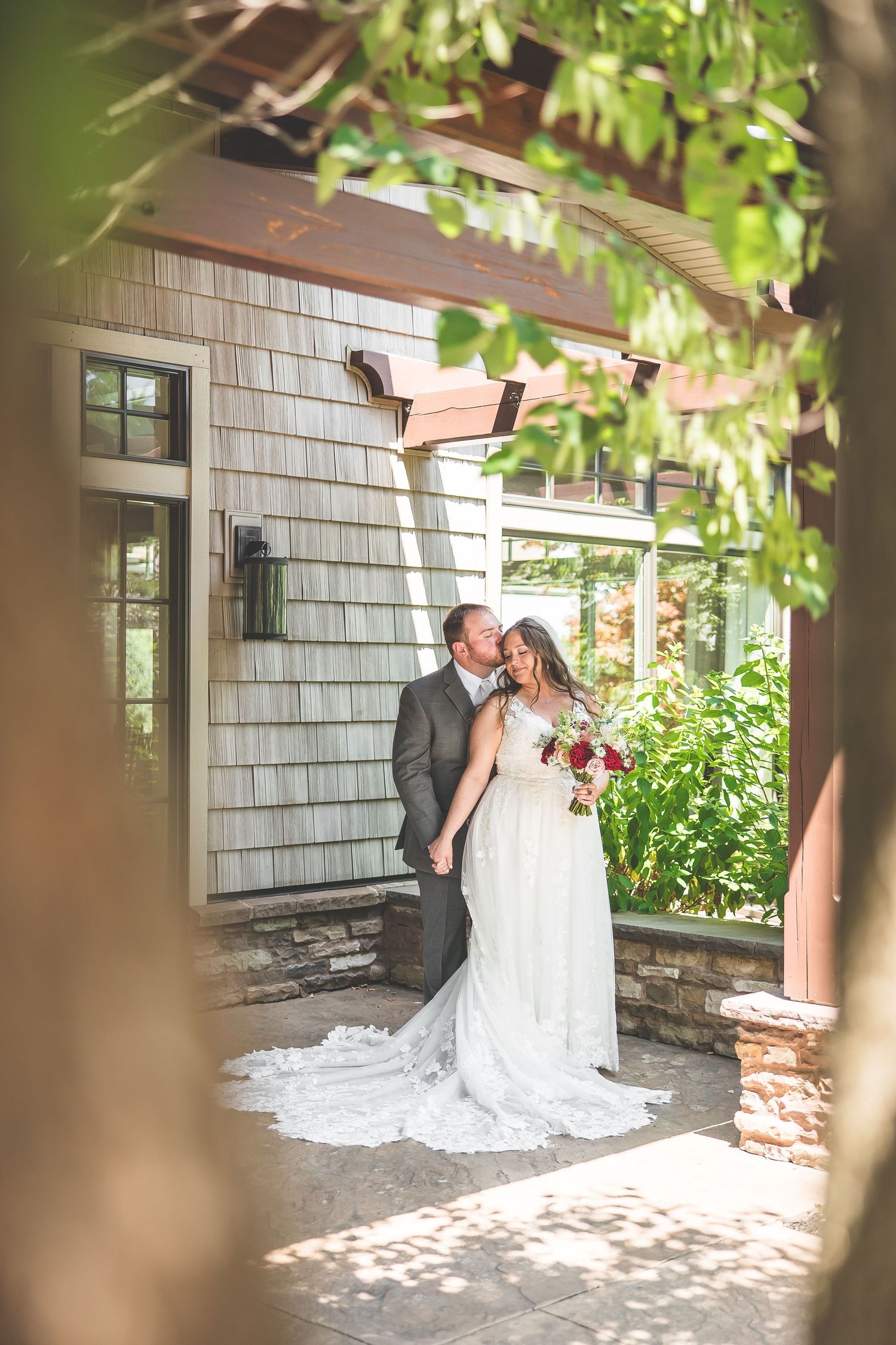 Romantic moment captured outside a white shingled house with green foliage overhead.