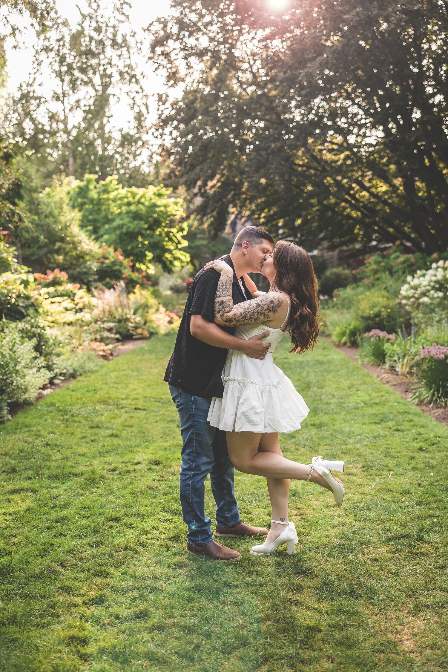 A romantic couple embraces in a garden setting with sunlight filtering through trees.