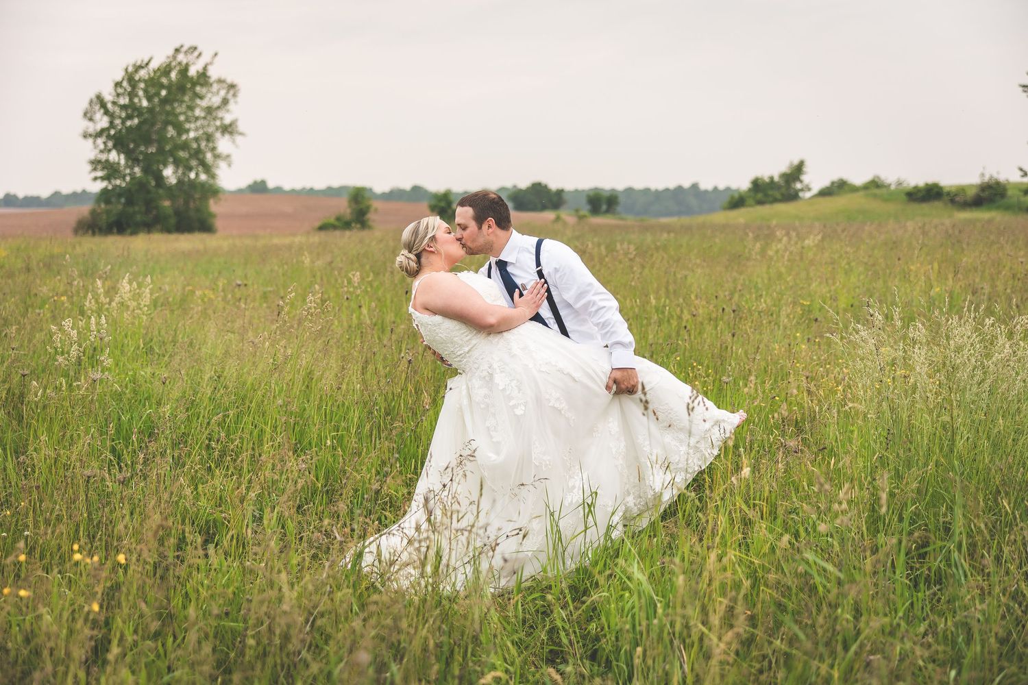 Wedding couple strolls through tall grass in a rustic countryside setting.