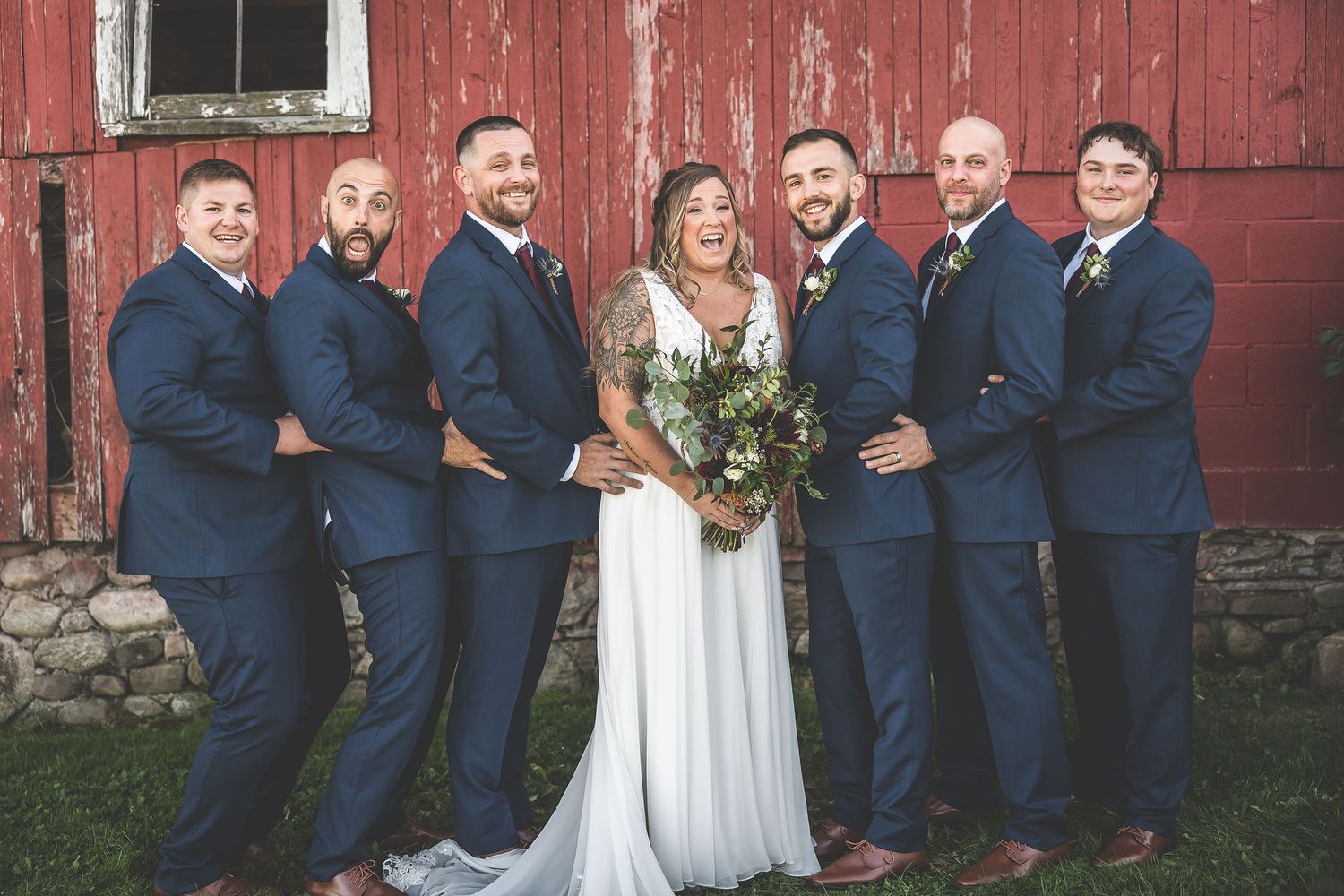 Groomsmen in navy suits pose together against rustic red barn backdrop.