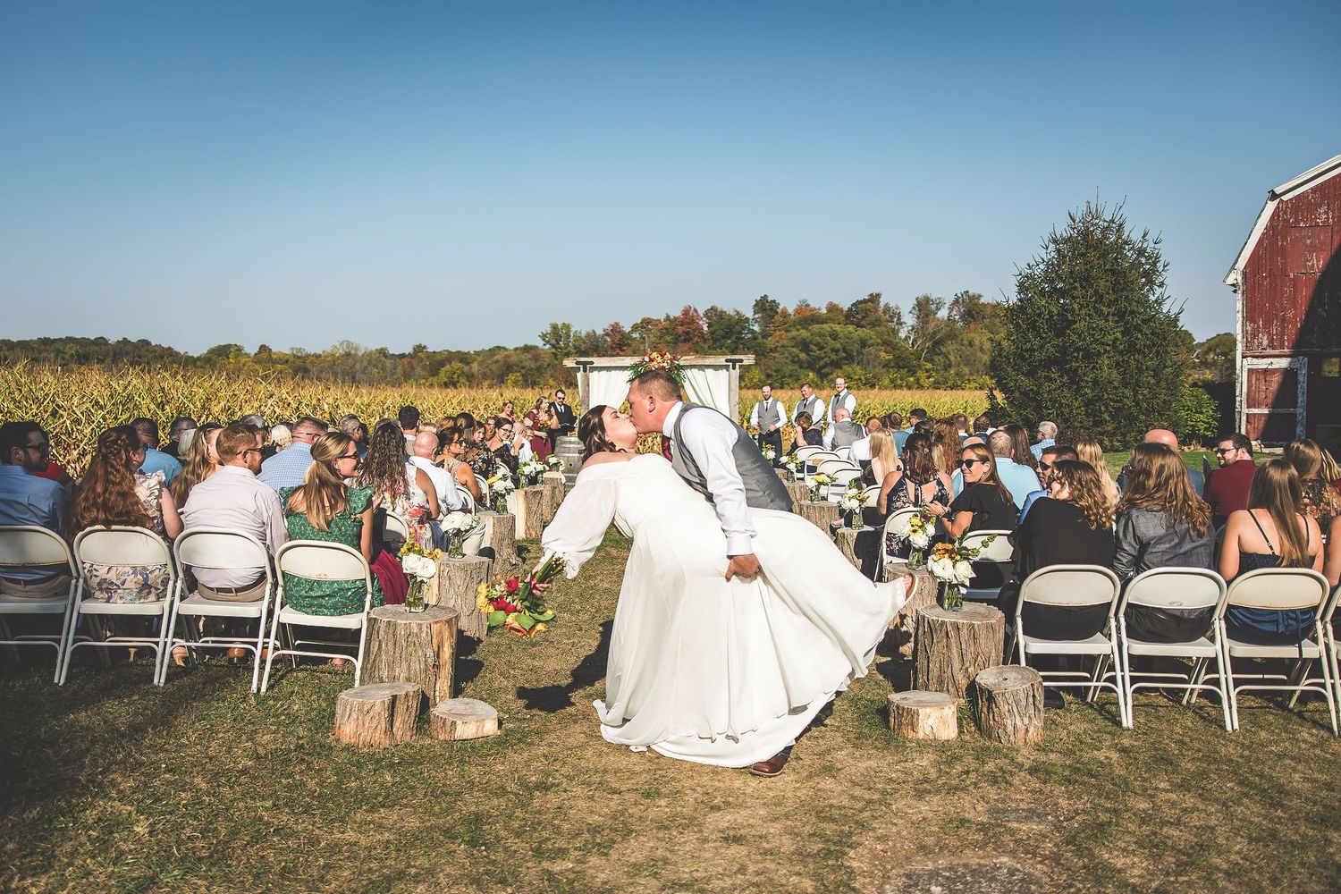 Wide angle view of outdoor wedding ceremony with guests seated on white chairs near a rustic barn.