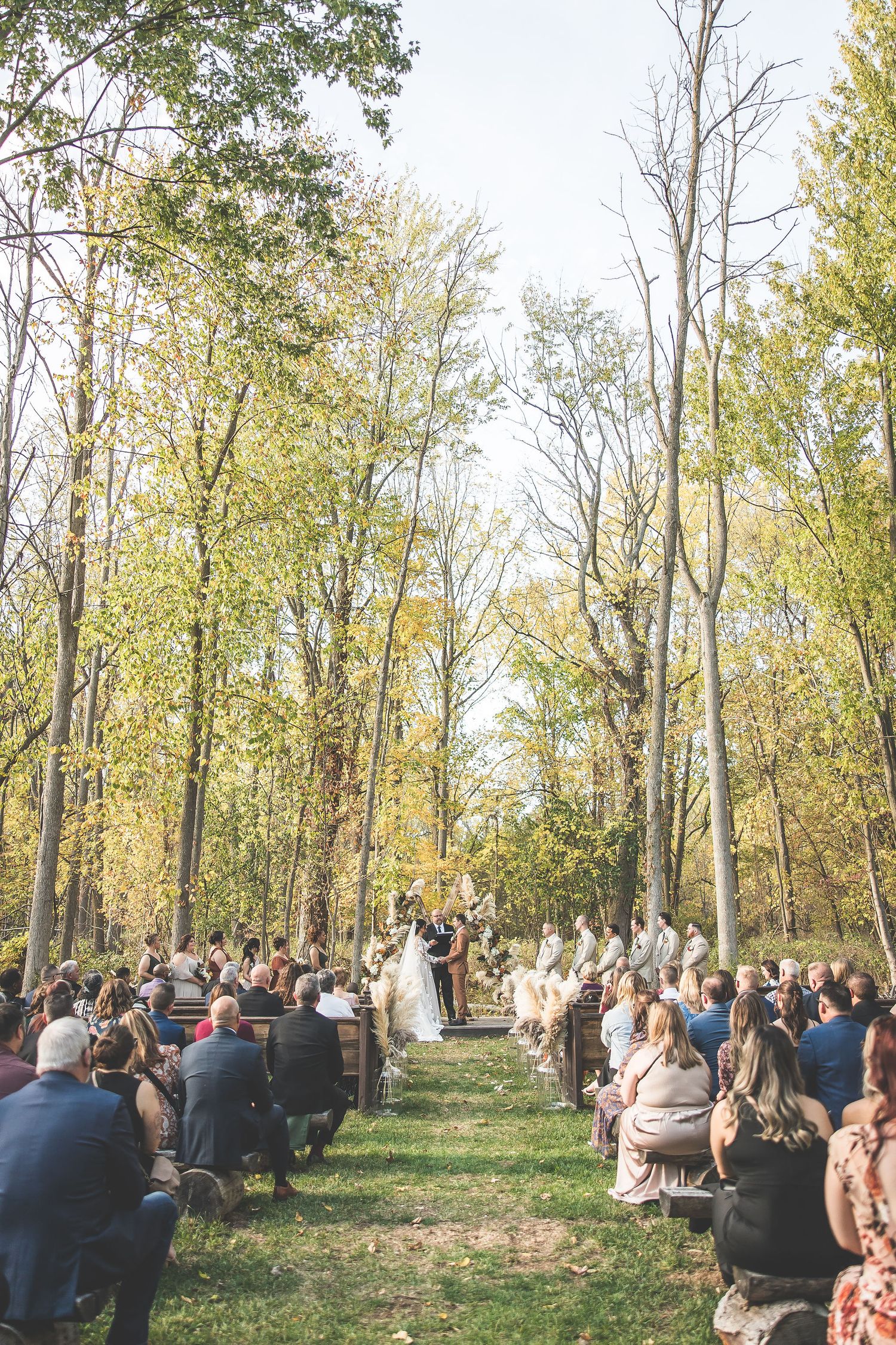 Wide view of forest wedding ceremony with guests seated among fall foliage.