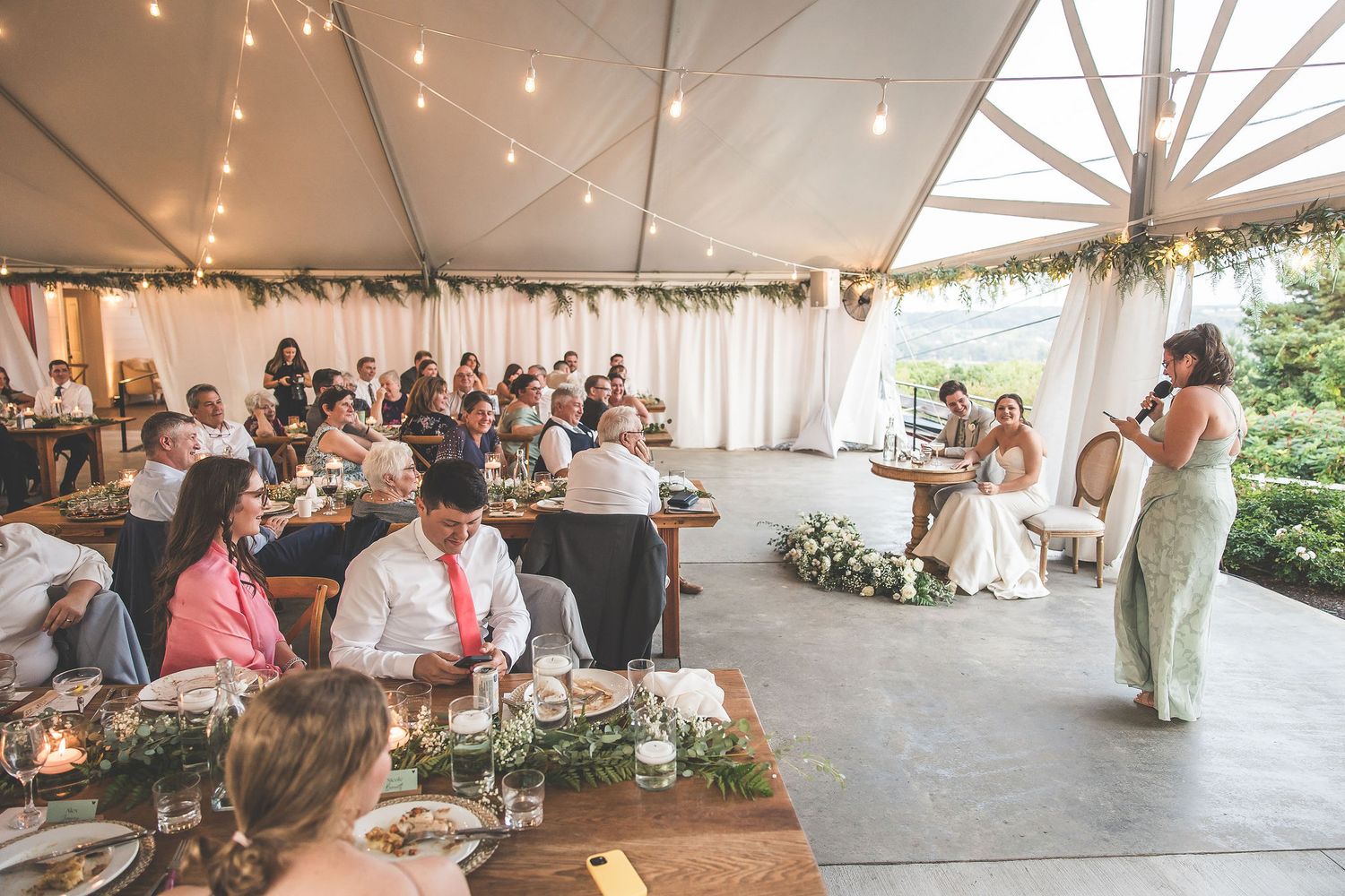 Wide view of a wedding reception under a tent with string lights and floral decorations.