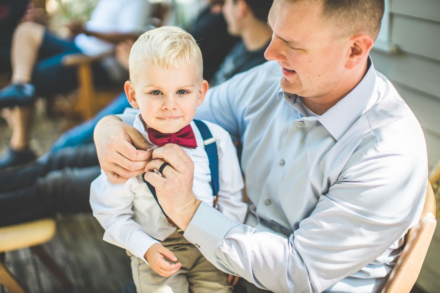 Young ring bearer in formal attire sits with adults during outdoor wedding ceremony.