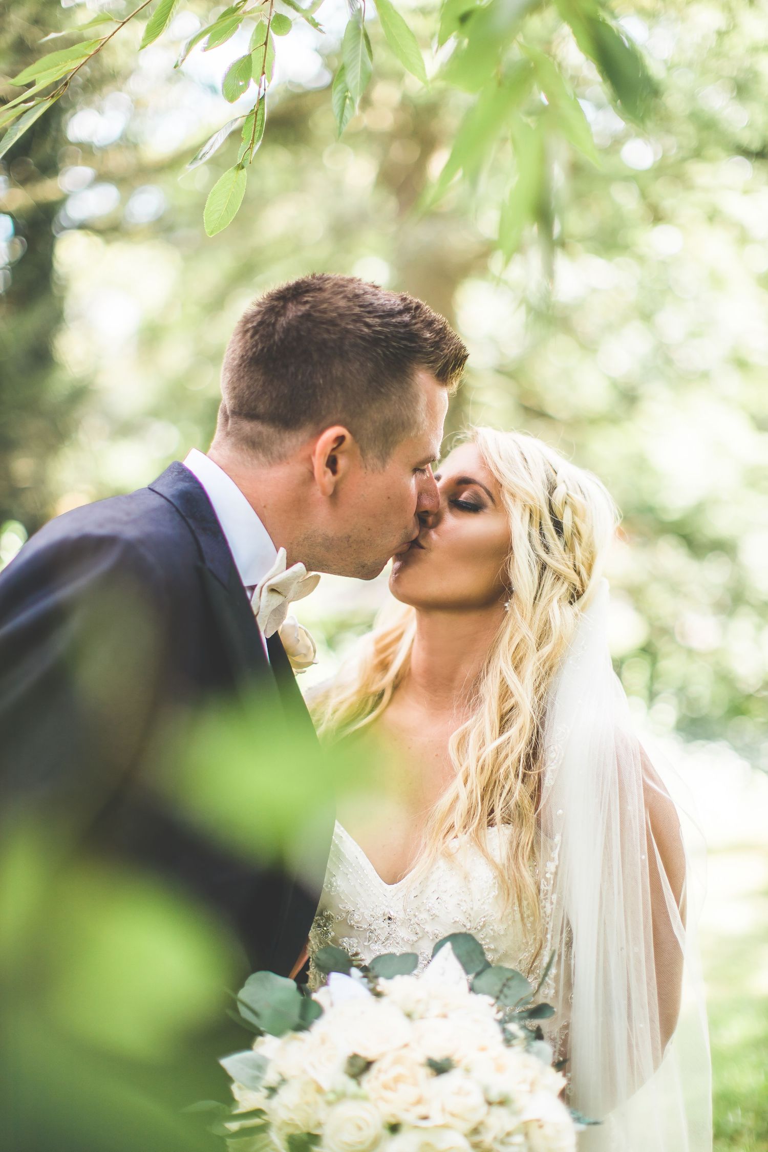 A romantic wedding portrait captured through green foliage with soft natural lighting.