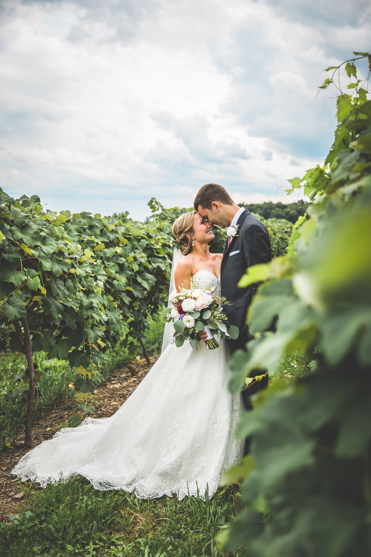 A romantic photoshoot in a lush vineyard with grapevines and cloudy skies in the background.