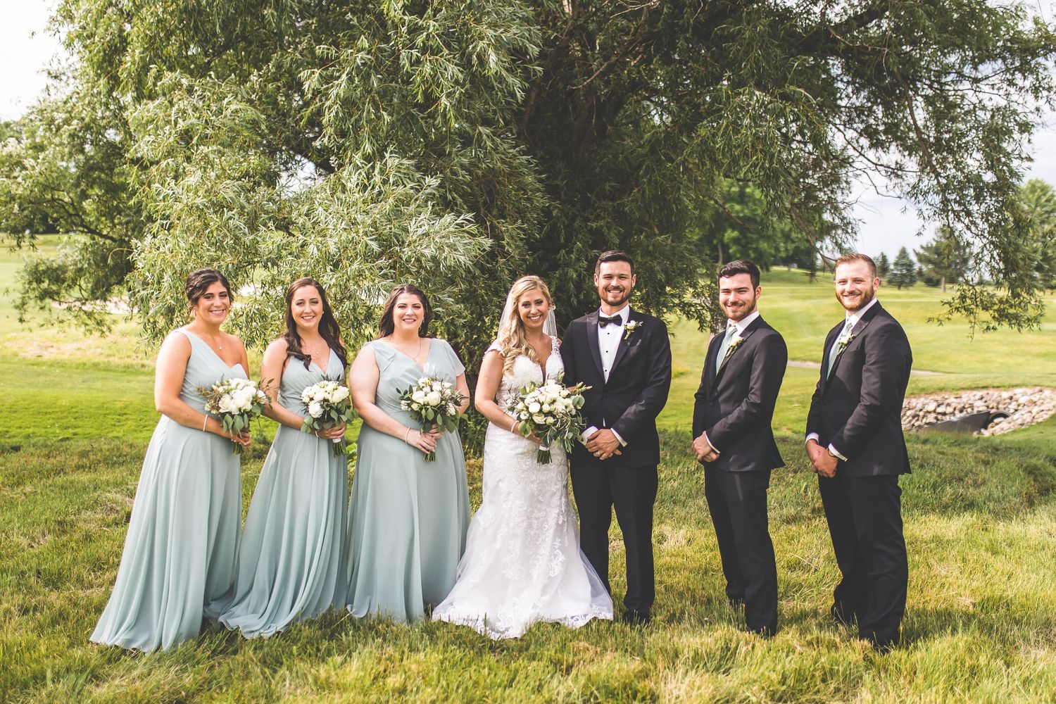 Wedding party in light blue dresses and dark suits posed outdoors under trees.