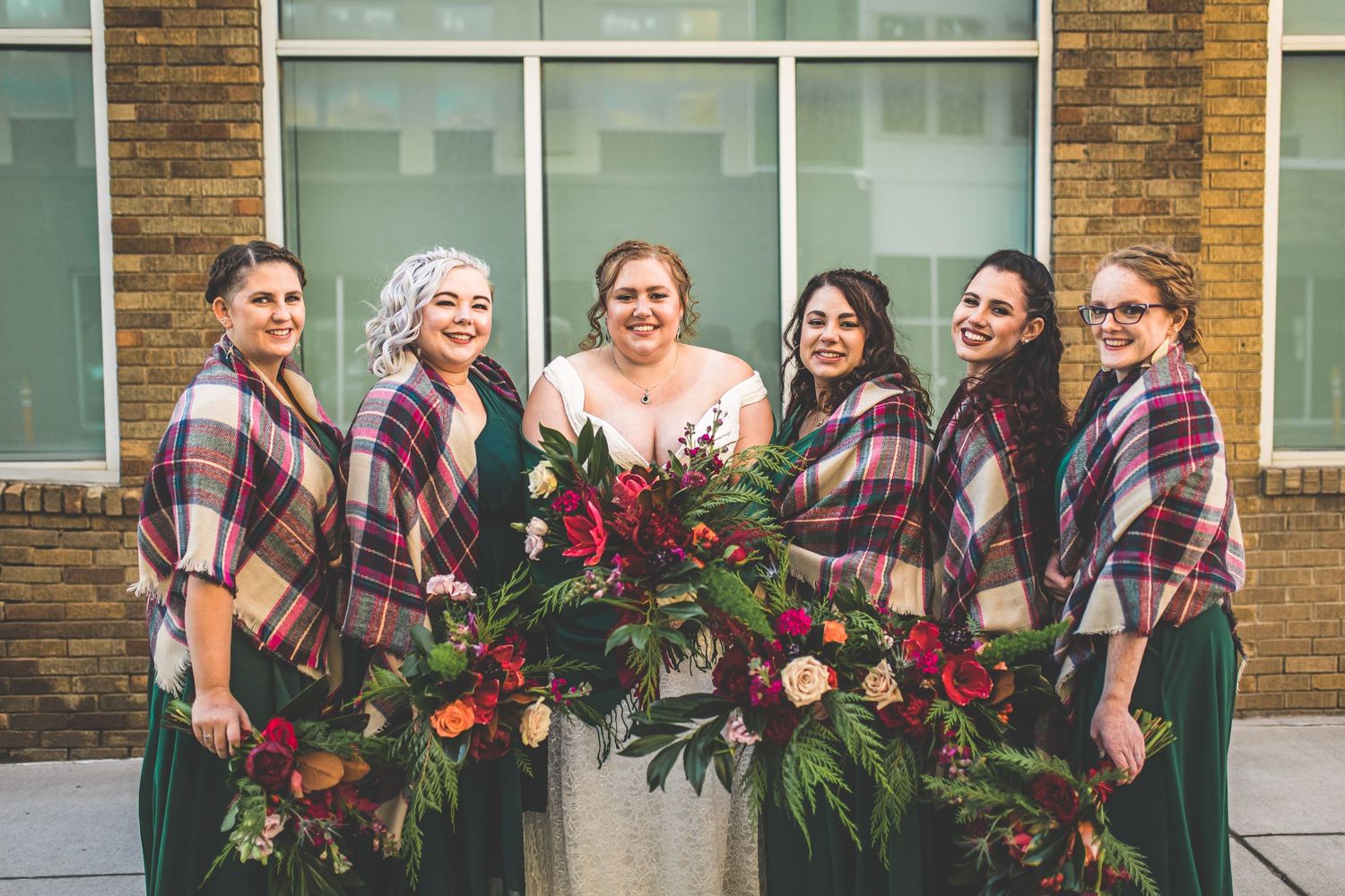Wedding party wearing plaid flannel shirts posing with burgundy floral arrangements.