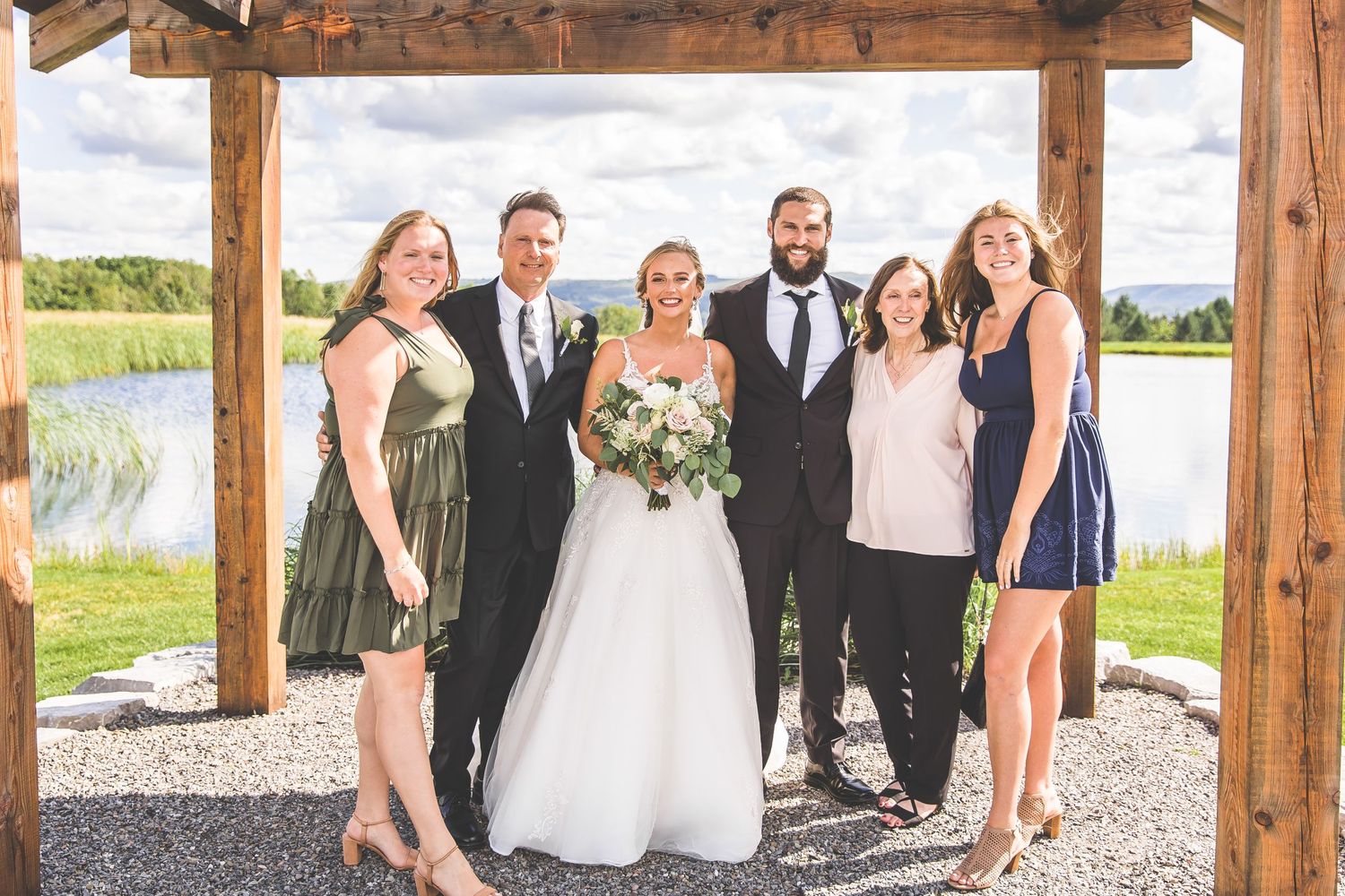 Wedding party poses outdoors under rustic wooden beams with lake and greenery in background.