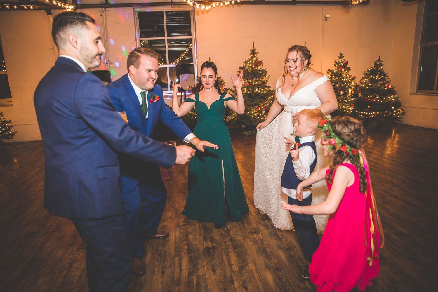 Wedding guests dancing and celebrating in a festively decorated reception hall with Christmas trees.