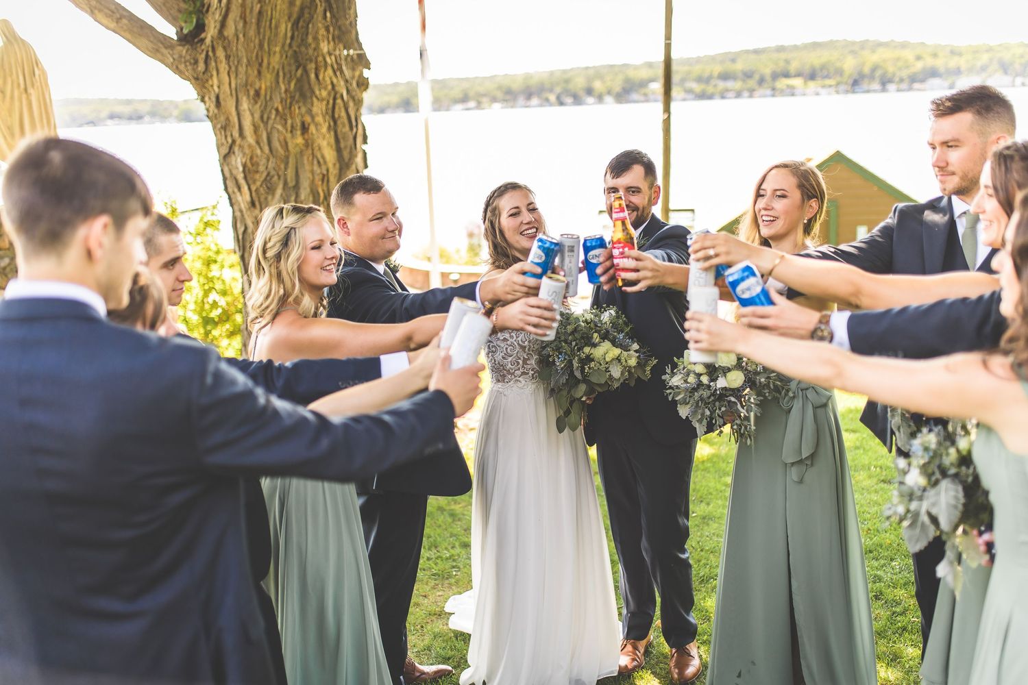 Wedding party toasts with drinks during outdoor lakeside celebration.