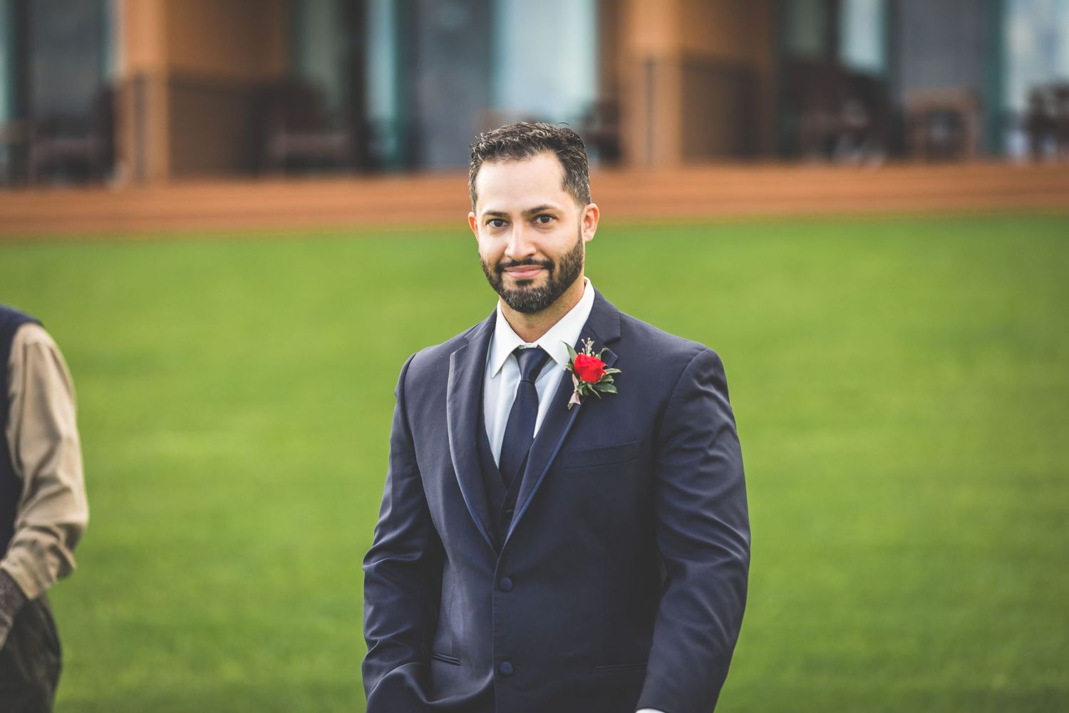 Outdoor portrait of a wedding guest in a navy suit with boutonniere against green lawn.