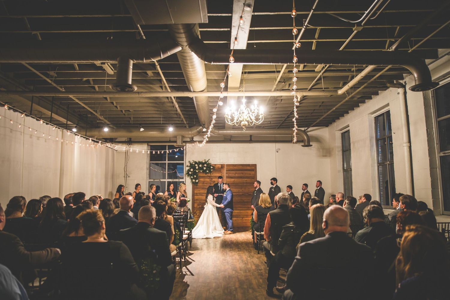 Wide angle view of wedding reception venue with exposed ceiling and string lights.