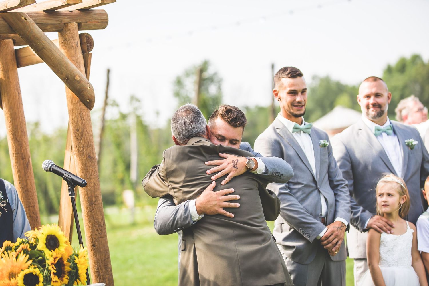 Rustic wooden arbor decorated with sunflowers frames an outdoor wedding ceremony.