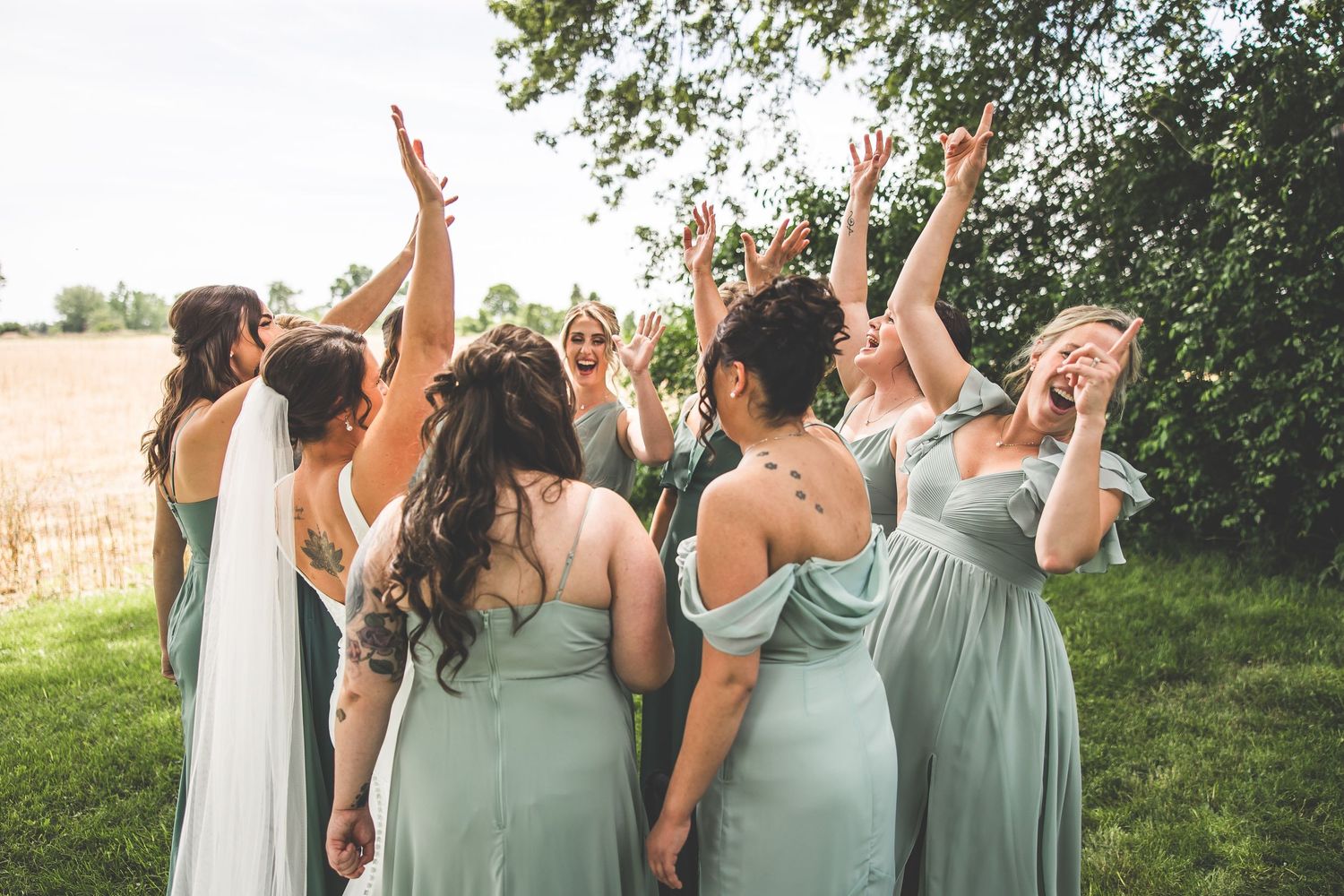 Bridesmaids in seafoam dresses celebrating with raised arms on a sunny lawn.