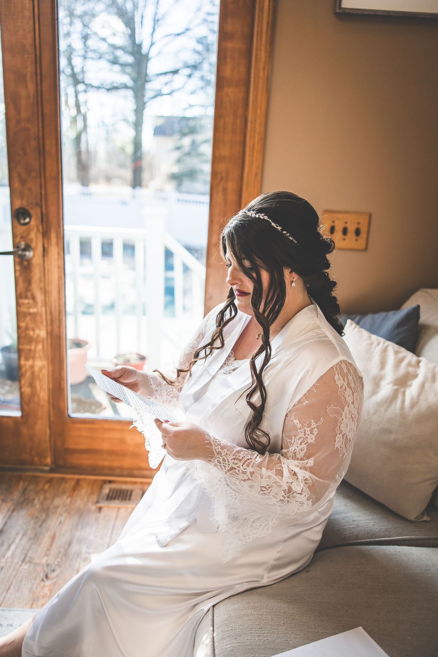 Bride in white lace robe relaxing by a window during wedding morning preparations.