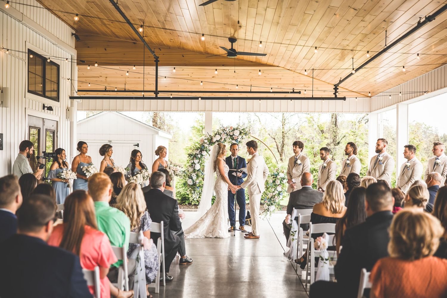 Wide view of wedding ceremony in modern venue with wooden ceiling and floral arch.