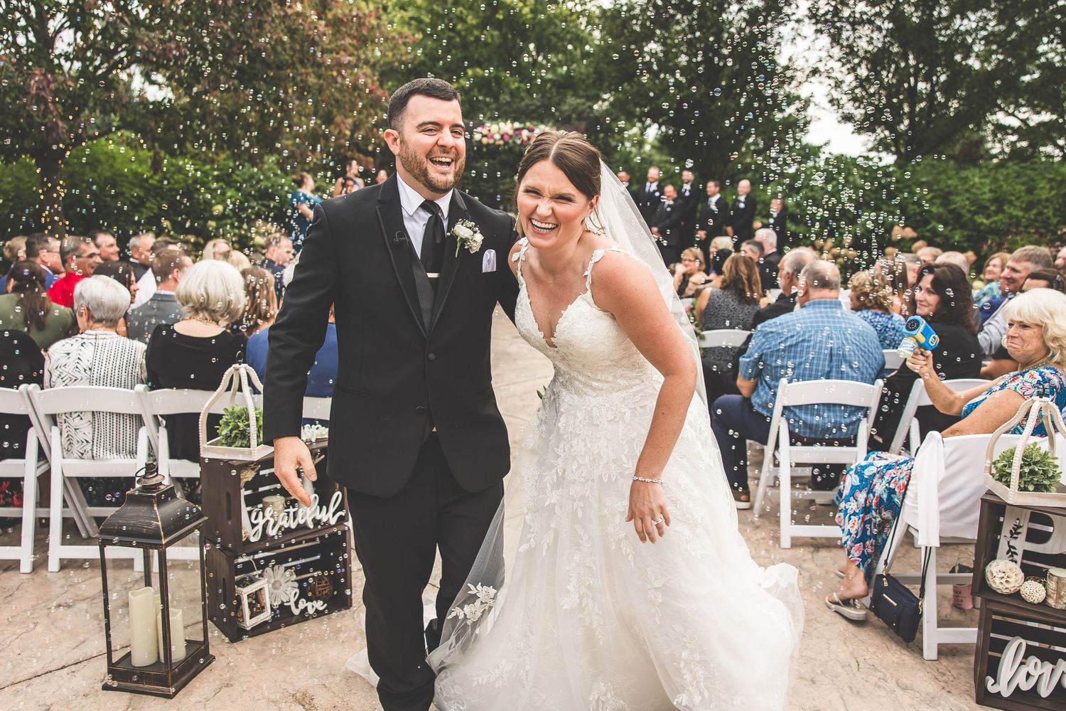 Wedding celebration with guests cheering as couple walks down outdoor aisle.