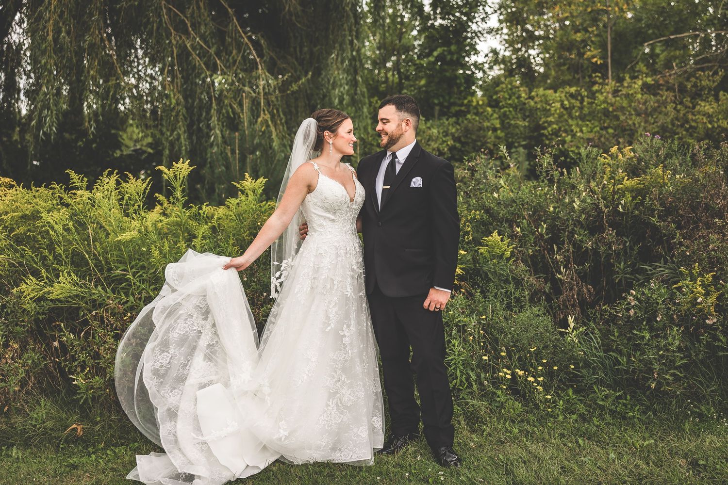 A bride's white dress flows beautifully as she walks through a sunlit garden with her groom.
