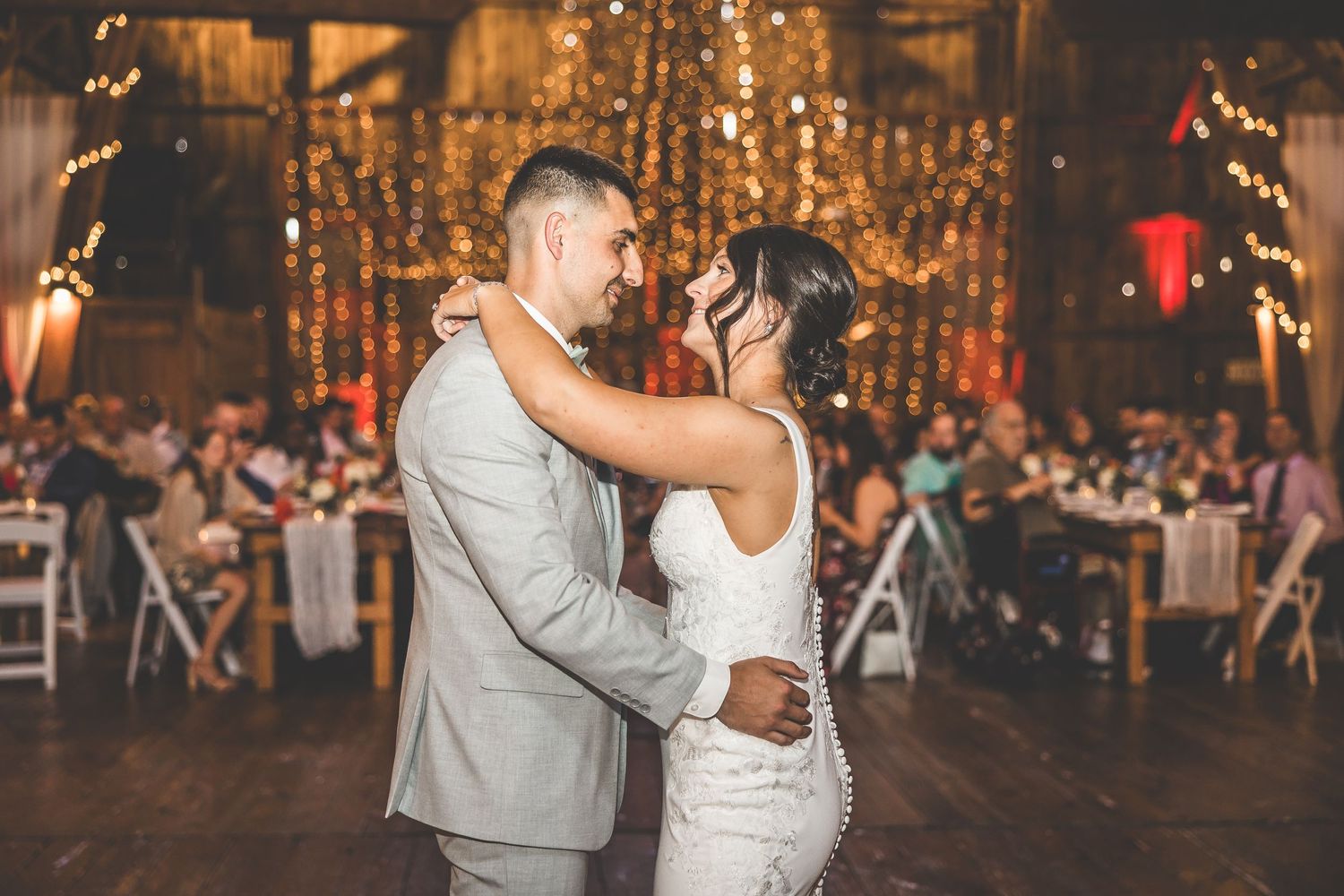 A romantic dance moment captured in a warmly lit barn reception with string lights and chandeliers.
