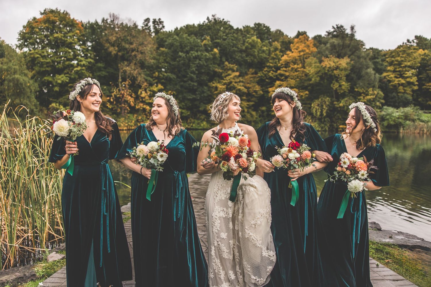 Bridal party laughs together in emerald velvet dresses by autumn lakeside.