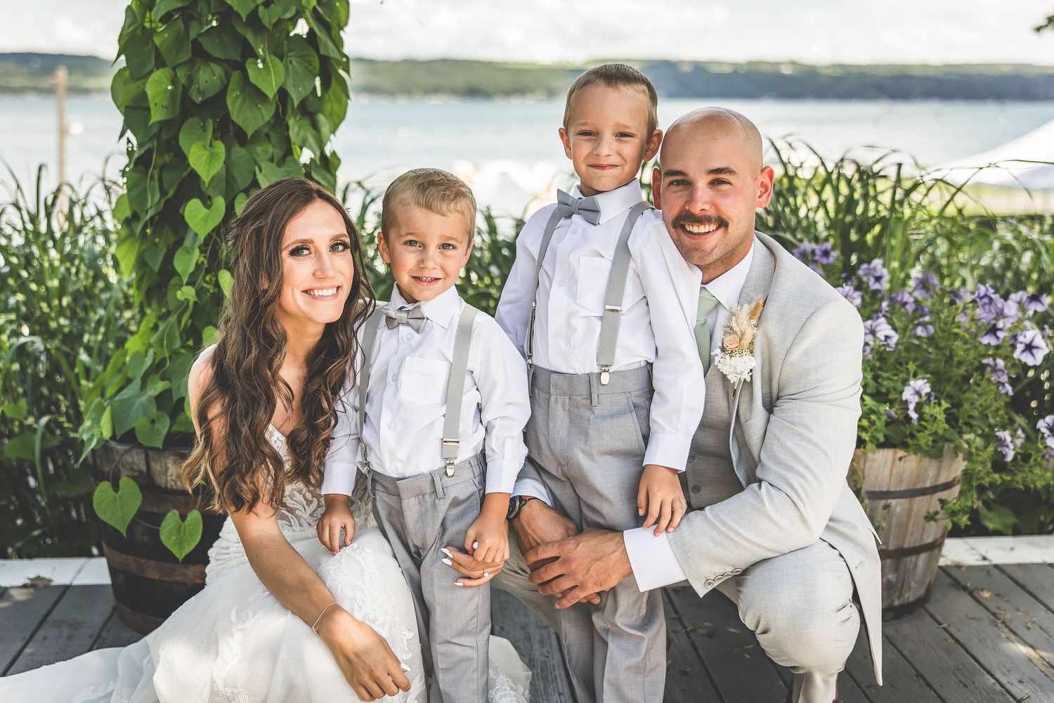 A wedding party in formal attire poses together on a waterfront deck with lush greenery.