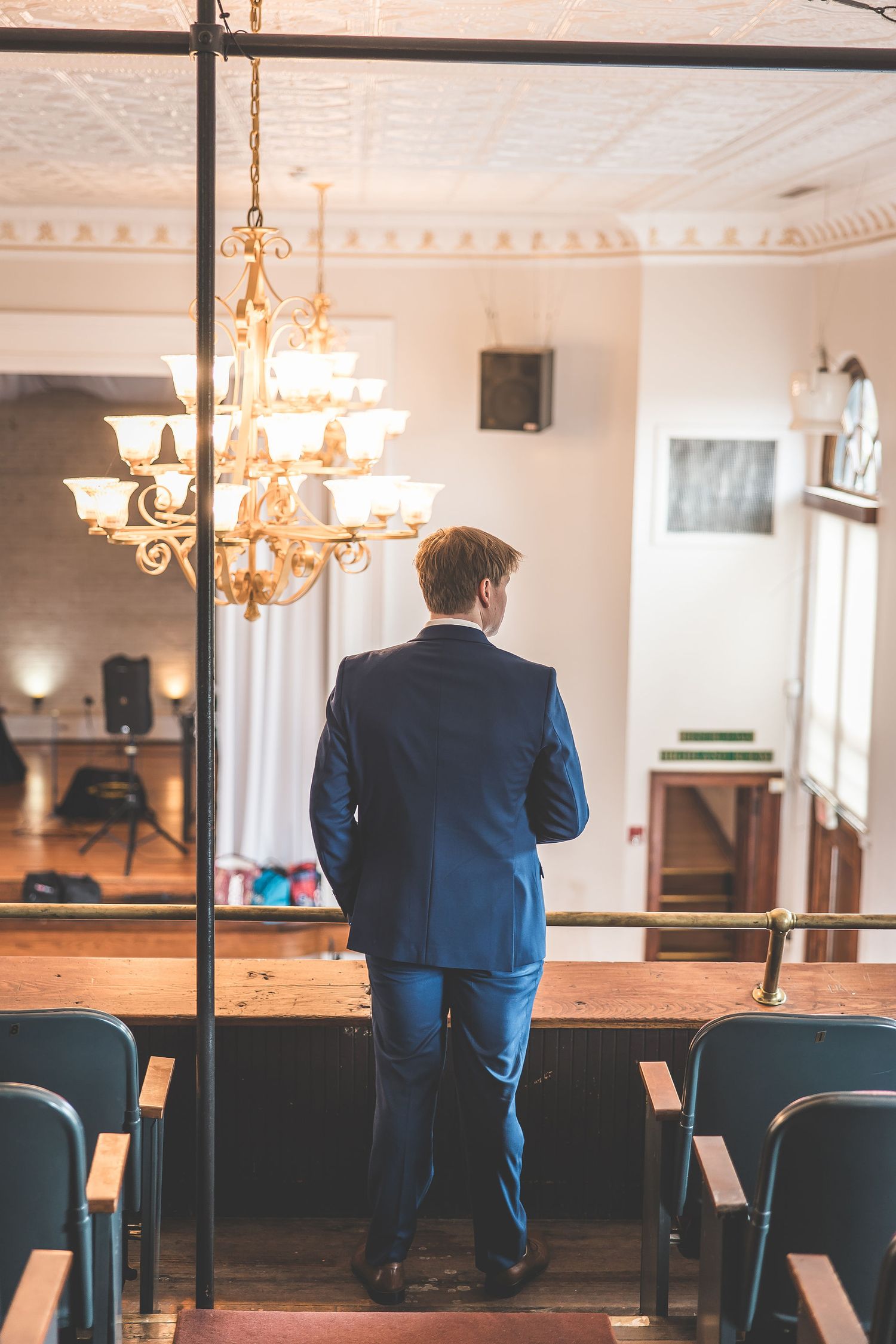 Person in a blue suit sits alone in an elegant venue with chandeliers and rows of chairs.
